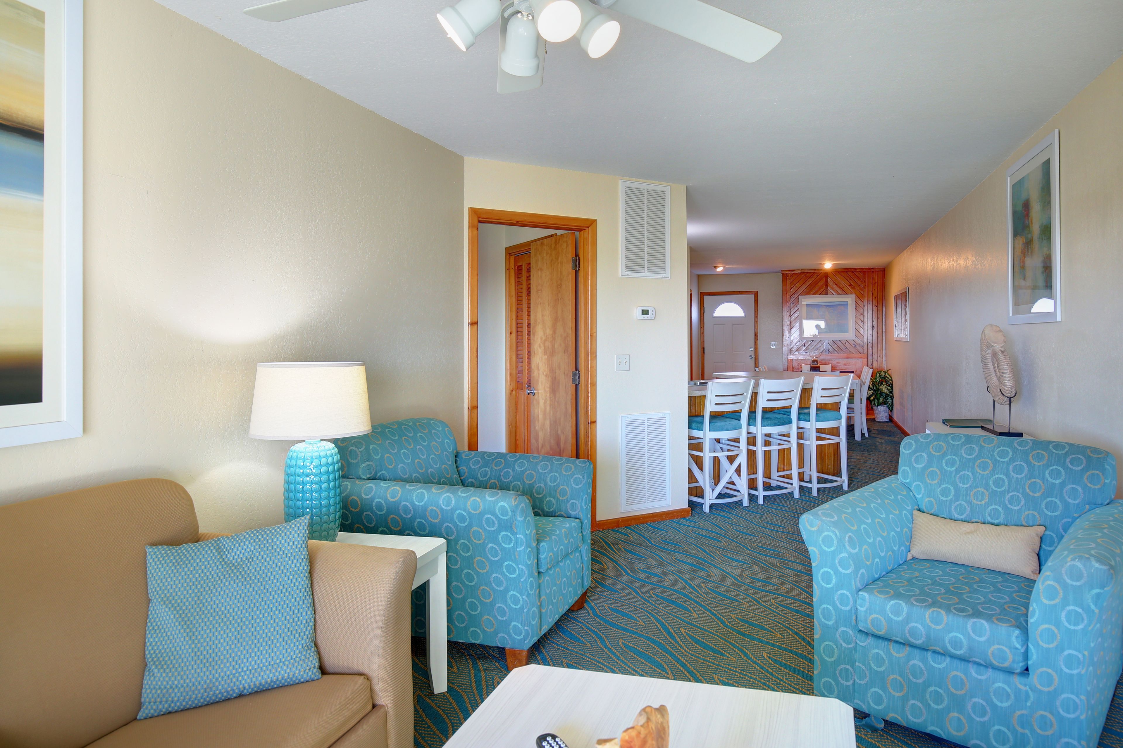 A room with two patterned sofas, a small table, and a ceiling fan. There is a door leading to a dining area with chairs and a table.