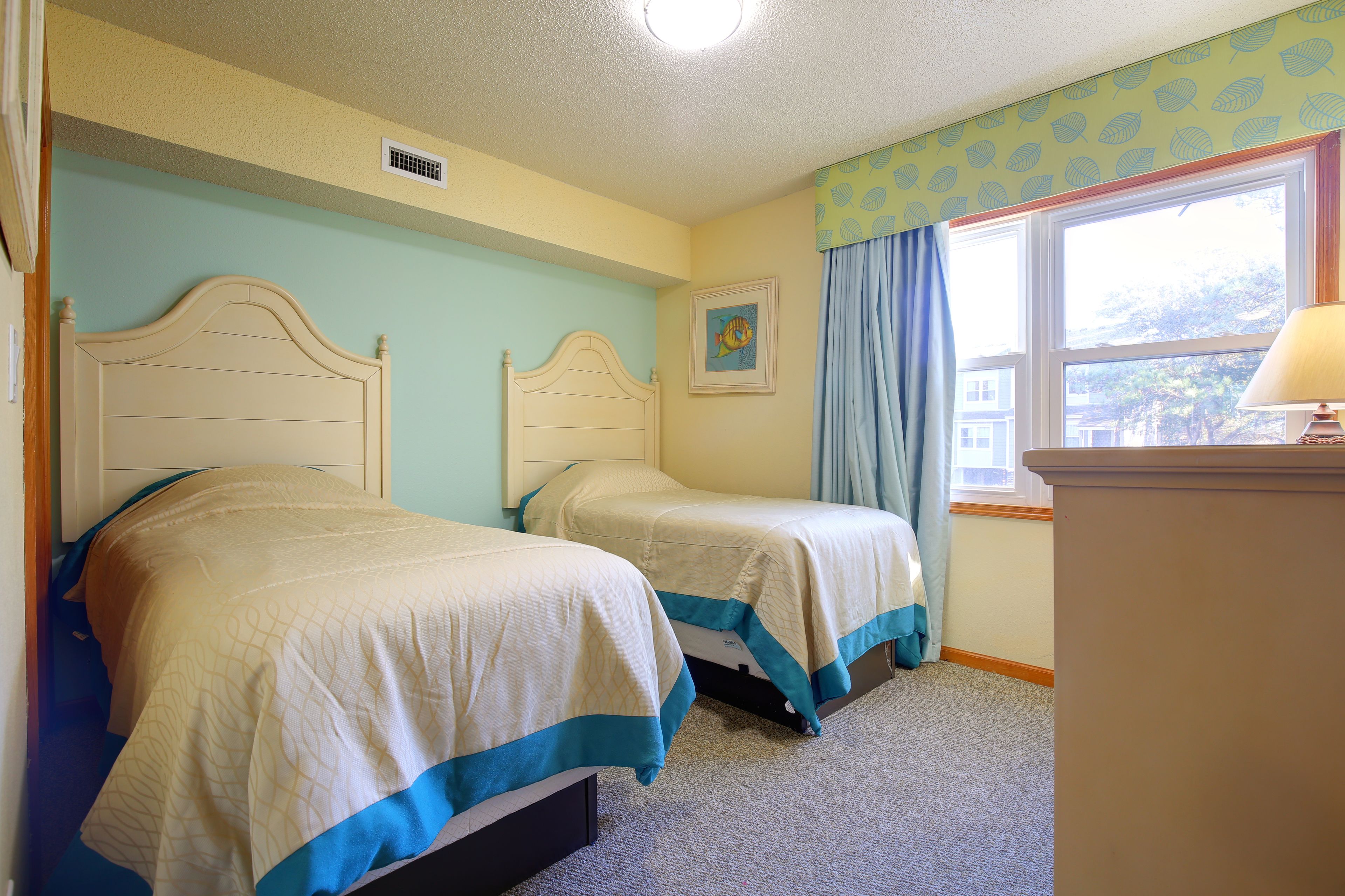 A bedroom with two beds, a window with curtains, a lamp, and a painting on the wall.