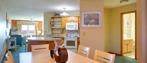 A compact kitchen with a microwave, stove, and wooden cabinets. There is a dining area with a table and chairs, and a small seating area with a blue chair.