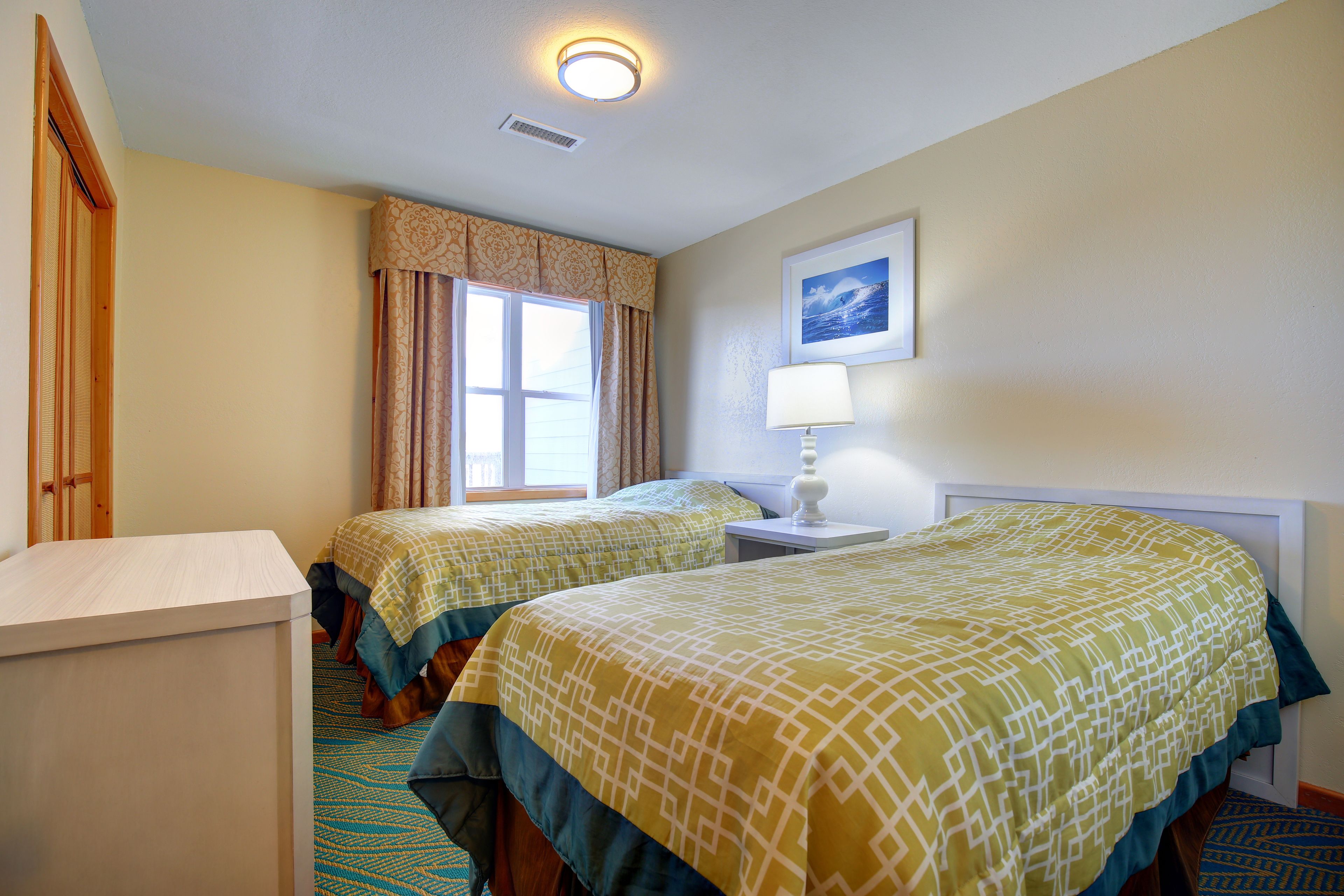 A hotel room with two beds, a desk, a lamp, a window with curtains, and a framed picture on the wall.