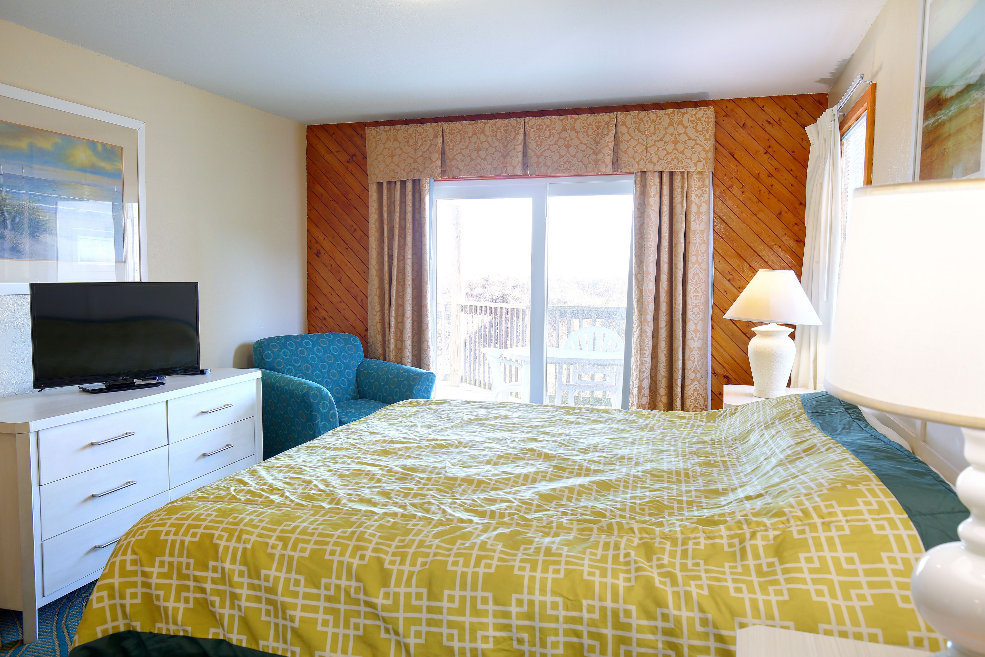 A hotel room with a bed, a TV on a dresser, a chair, a lamp, and a window with curtains.