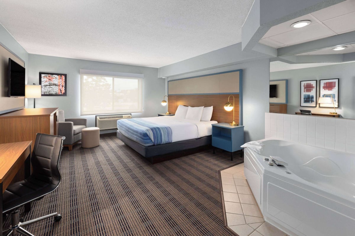 A modern hotel room with a large bed, a desk, a chair, a desk lamp, a nightstand, a bathtub, and a window.
