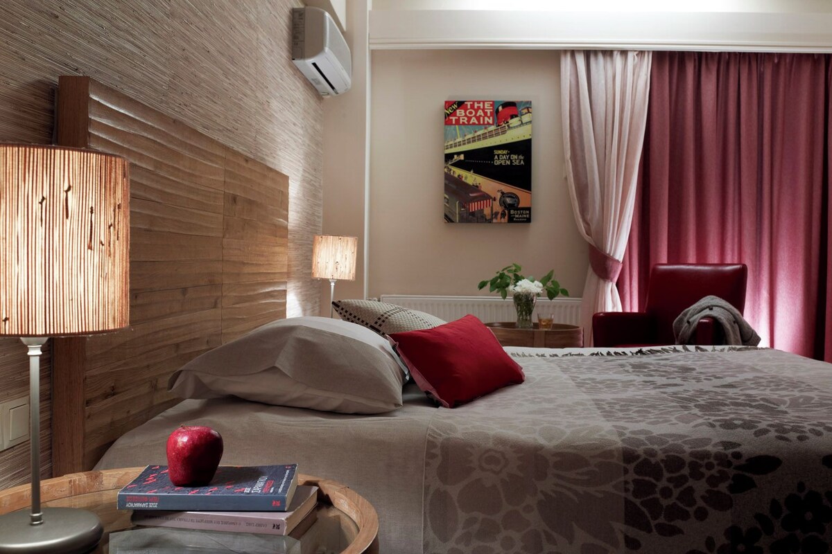 A hotel room with a wooden headboard, a bed with a floral-patterned bedspread, a red pillow, a bedside table with a lamp, a vase with flowers, and a poster on the wall.