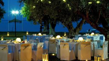 An outdoor dining area with tables set for a meal, surrounded by trees and a view of the sea.