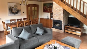 Una sala de estar con un sofá gris, una mesa de comedor de madera y sillas. Una escalera de madera conduce a un nivel superior.