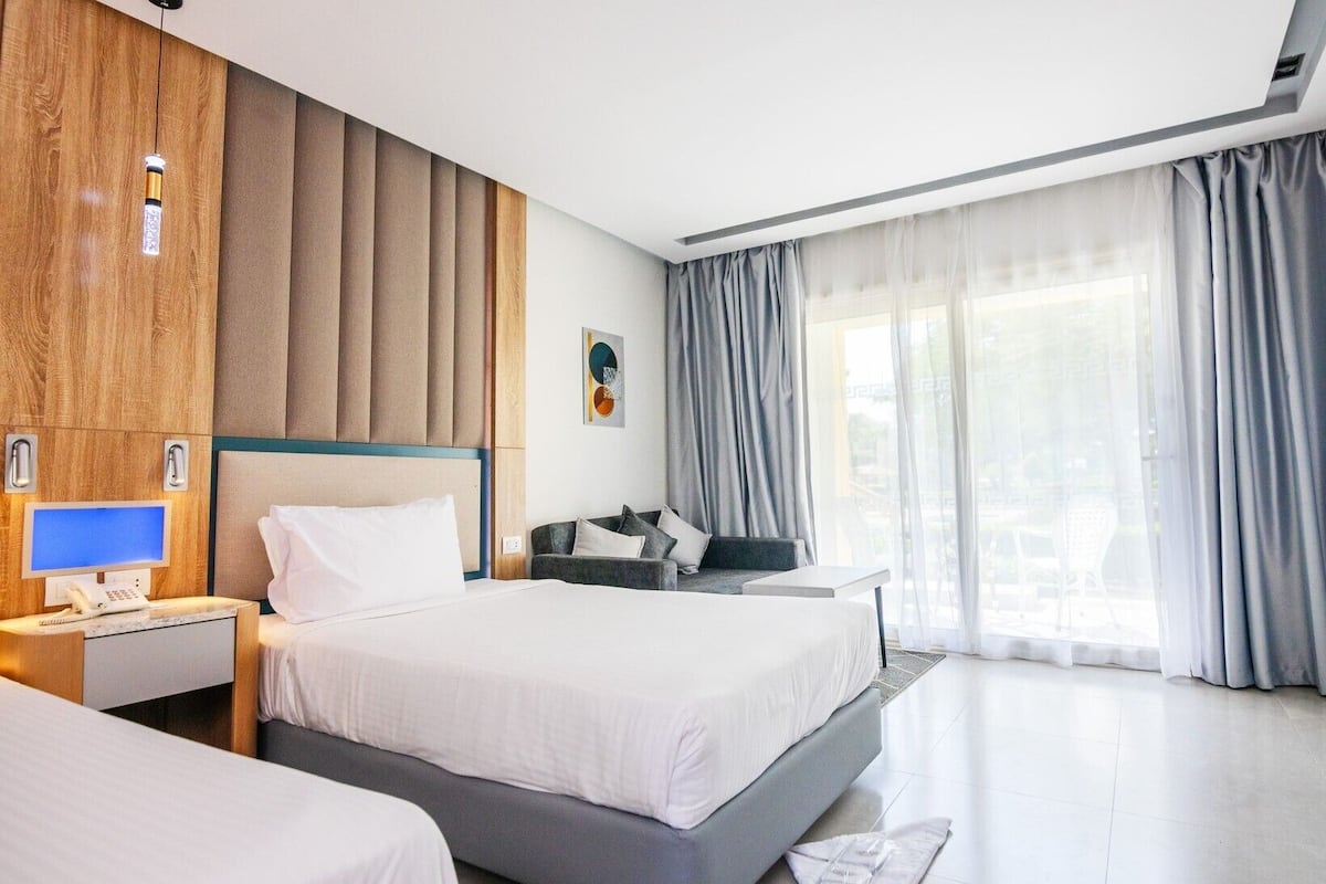 Quarto de hotel com uma cama grande, uma escrivaninha com computador, uma cadeira e uma janela com cortinas.