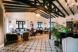 A spacious living room with a high ceiling, exposed beams, and a chandelier. There are multiple seating areas with sofas and chairs, a large potted plant, and a patterned floor.