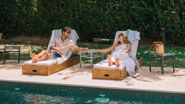Two lounge chairs by a pool with a wooden deck, surrounded by greenery and a stone wall.