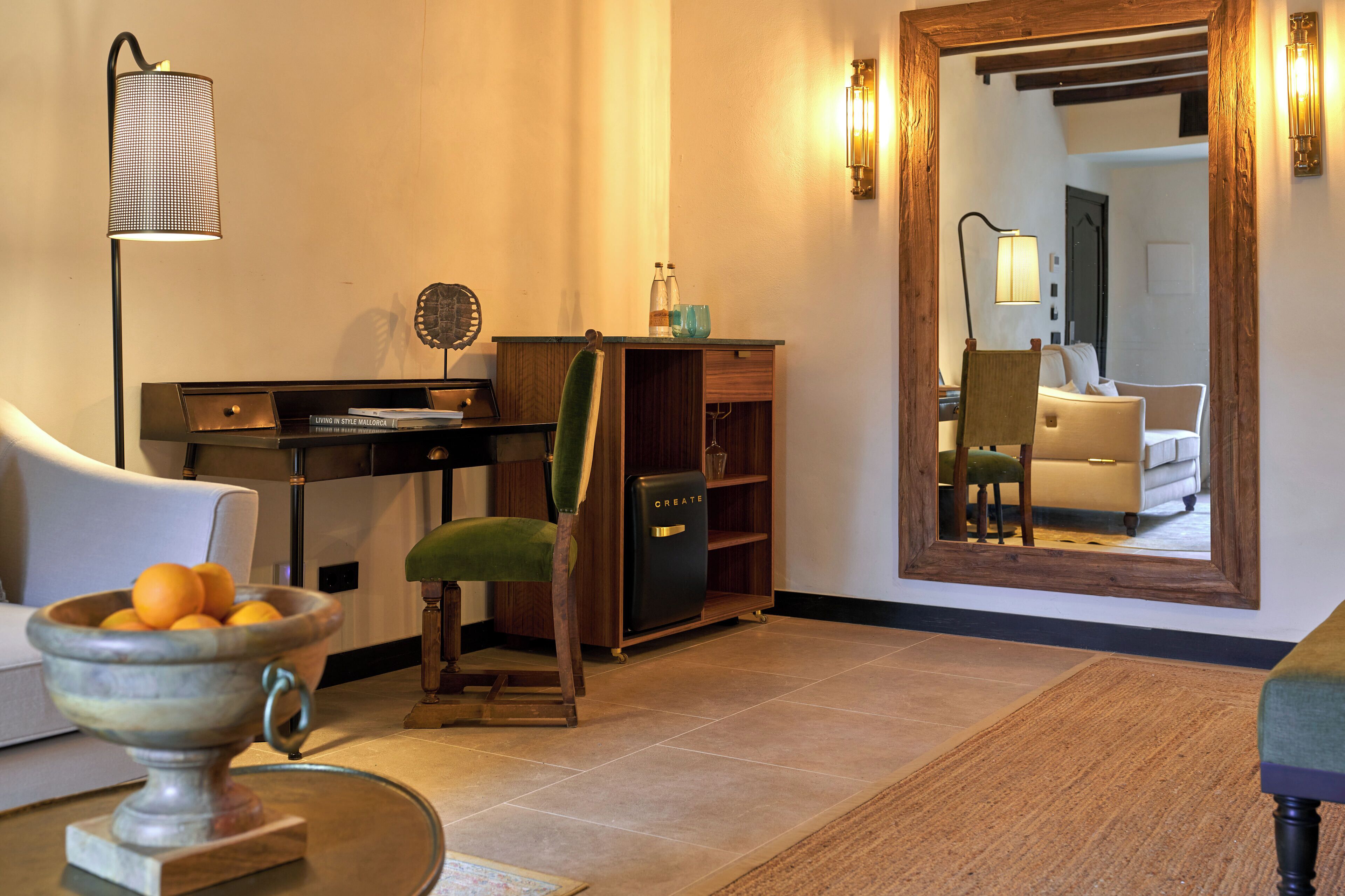 A living room with a sofa, a chair, a coffee table, a lamp, a bookshelf, and a large mirror reflecting the room.