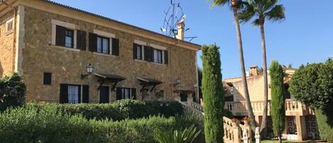 A stone building with multiple windows and a well-maintained garden with palm trees and hedges.