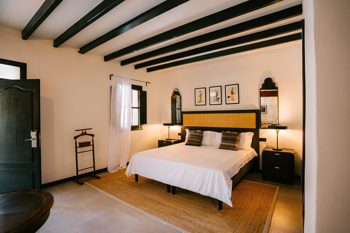 Een slaapkamer met een groot bed, houten balken in het plafond, een nachtkastje met een lamp en een klein houten krukje.