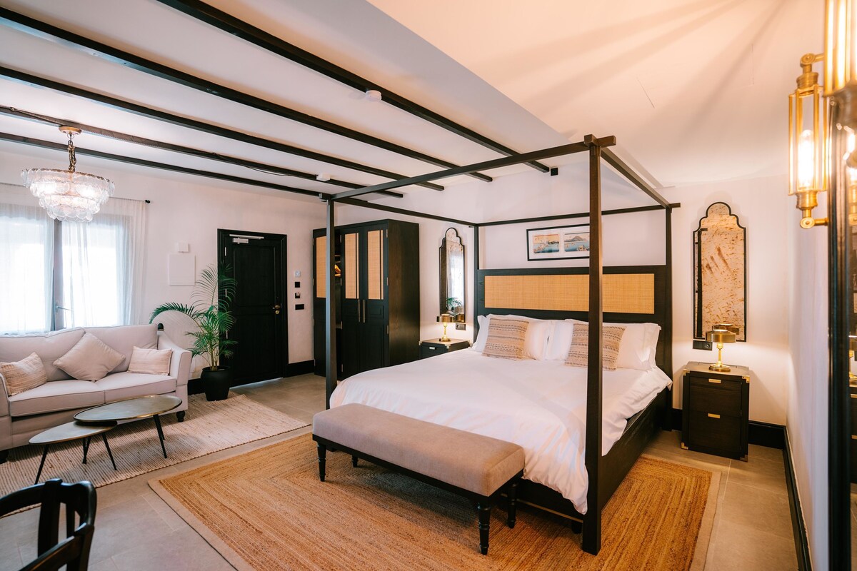 Een ruime slaapkamer met een hemelbed, een zithoek met een bank en salontafel, en een groot raam dat zorgt voor natuurlijk licht.