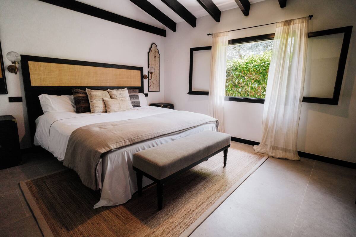 A bedroom with a large bed, a bench, a nightstand, a wall-mounted lamp, and a window with curtains.