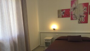 A bedroom with a bed, curtains, a nightstand, a wall-mounted light, and floral artwork. - Albergo Ester Di Fossi Laura (Rimini)