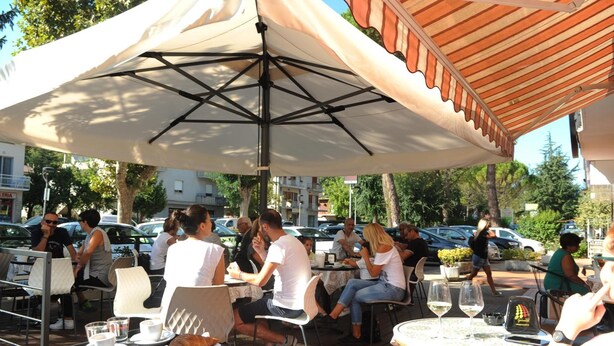 An outdoor café with tables and umbrellas, surrounded by parked cars and trees. - Albergo Ester di Fossi Laura (Rimini)