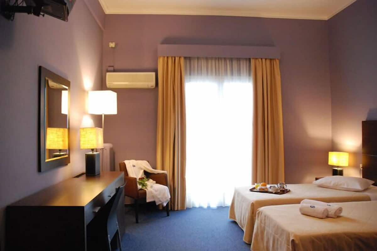 A hotel room with a desk, two beds, a chair, and a large window with curtains.