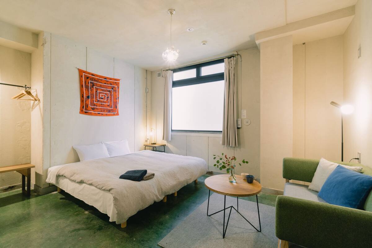 A hotel room with a bed, a green armchair, a small table, and a window with curtains.