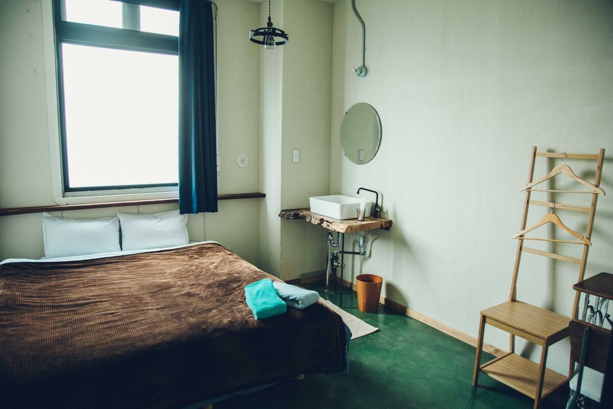 A bedroom with a bed, a window, a sink, a mirror, and a wooden chair.