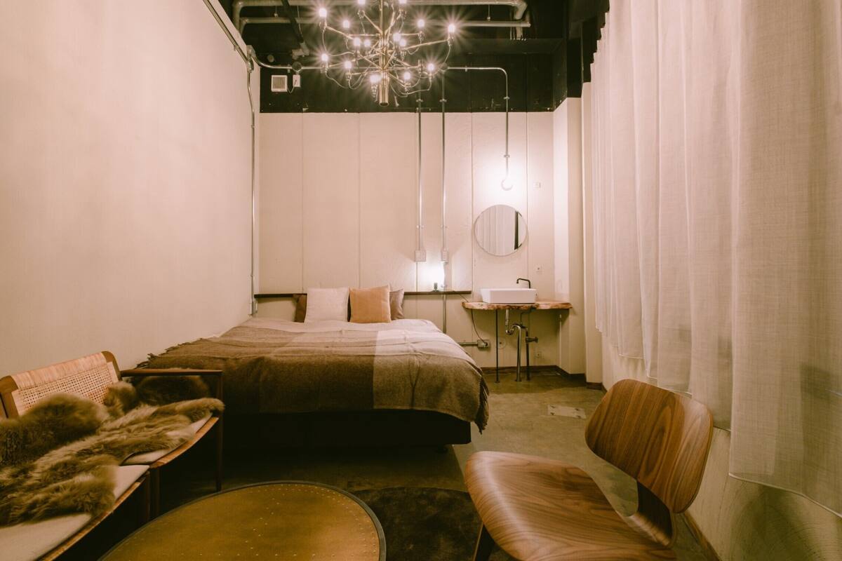 A bedroom with a bed, a chair, a round table, a mirror, and a sink.