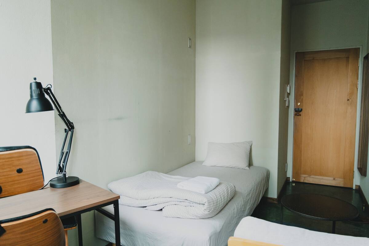 A single bed with a white duvet, a desk with a lamp, and a wooden door.