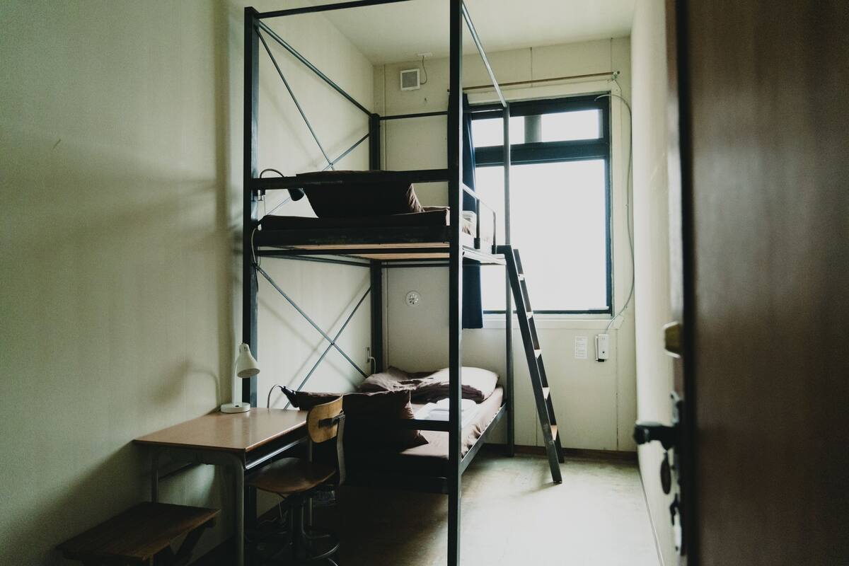 A dormitory room with bunk beds, a desk, a chair, and a window.