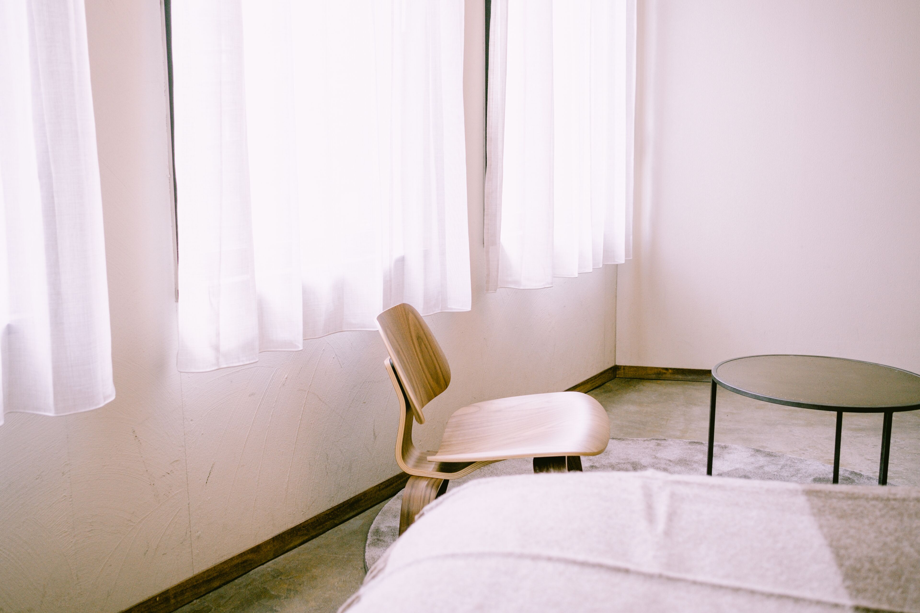 Une pièce avec une seule chaise et une petite table, une fenêtre ornée de rideaux légers et un mur sobre.
