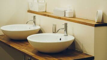 Two white sinks with chrome faucets on a wooden countertop, above which is a white shelf with various toiletries.