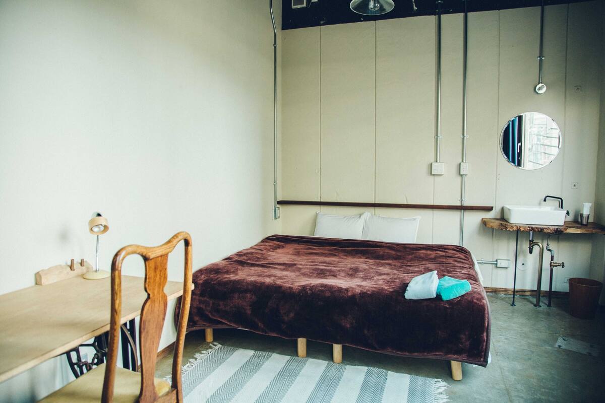 A bedroom with a bed, a desk, a chair, a mirror, and a small sink.