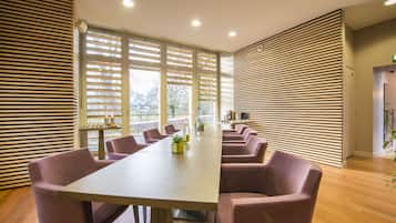 Une salle de conférence avec une longue table et plusieurs chaises, de grandes fenêtres équipées de stores et un mur en lambris de bois.