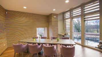 A dining area with a long table, six chairs, and a view of a pond through large windows.