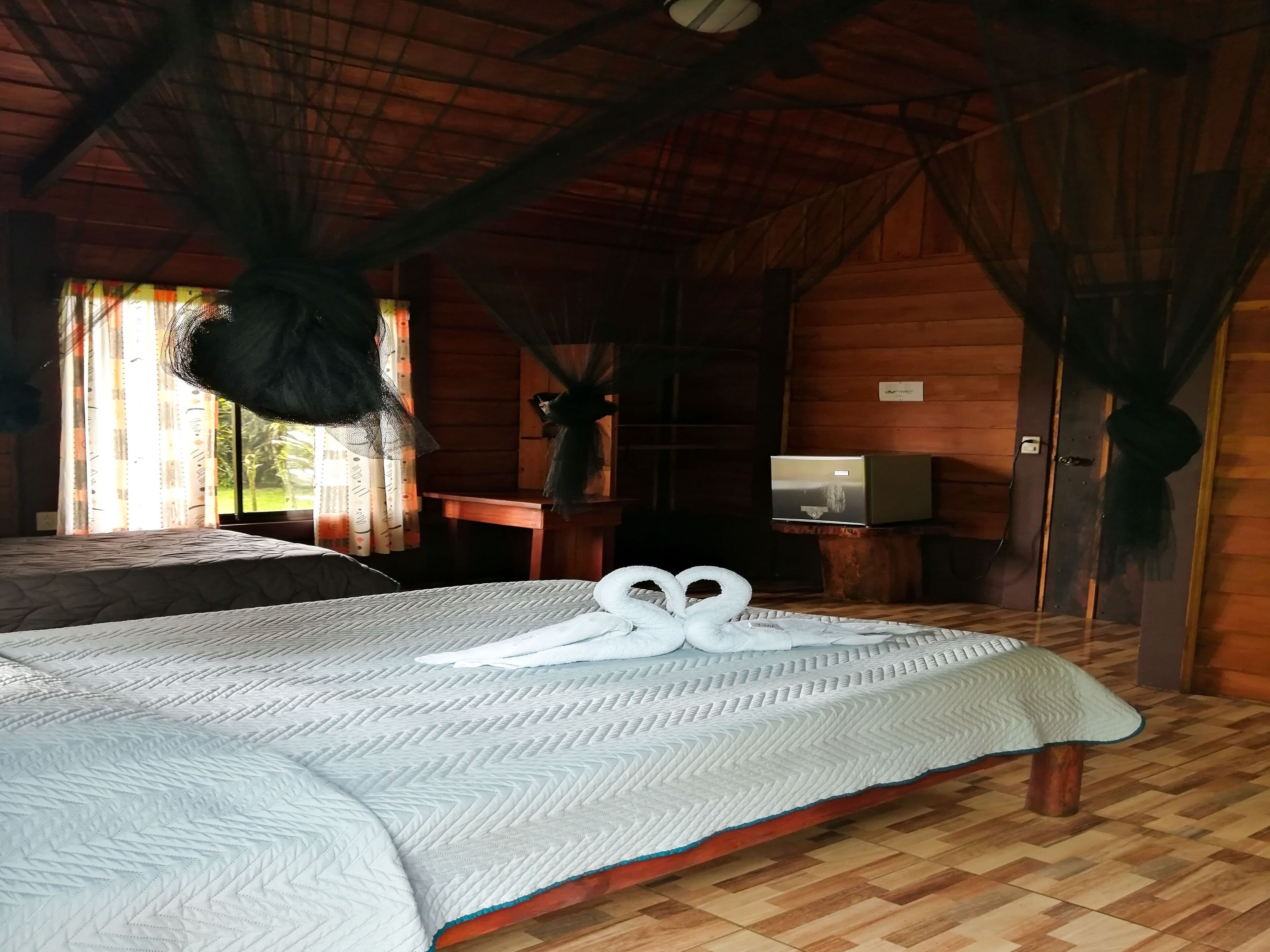 Quarto de cabana de madeira com uma cama, uma televisão sobre um suporte de madeira e uma janela com cortinas.