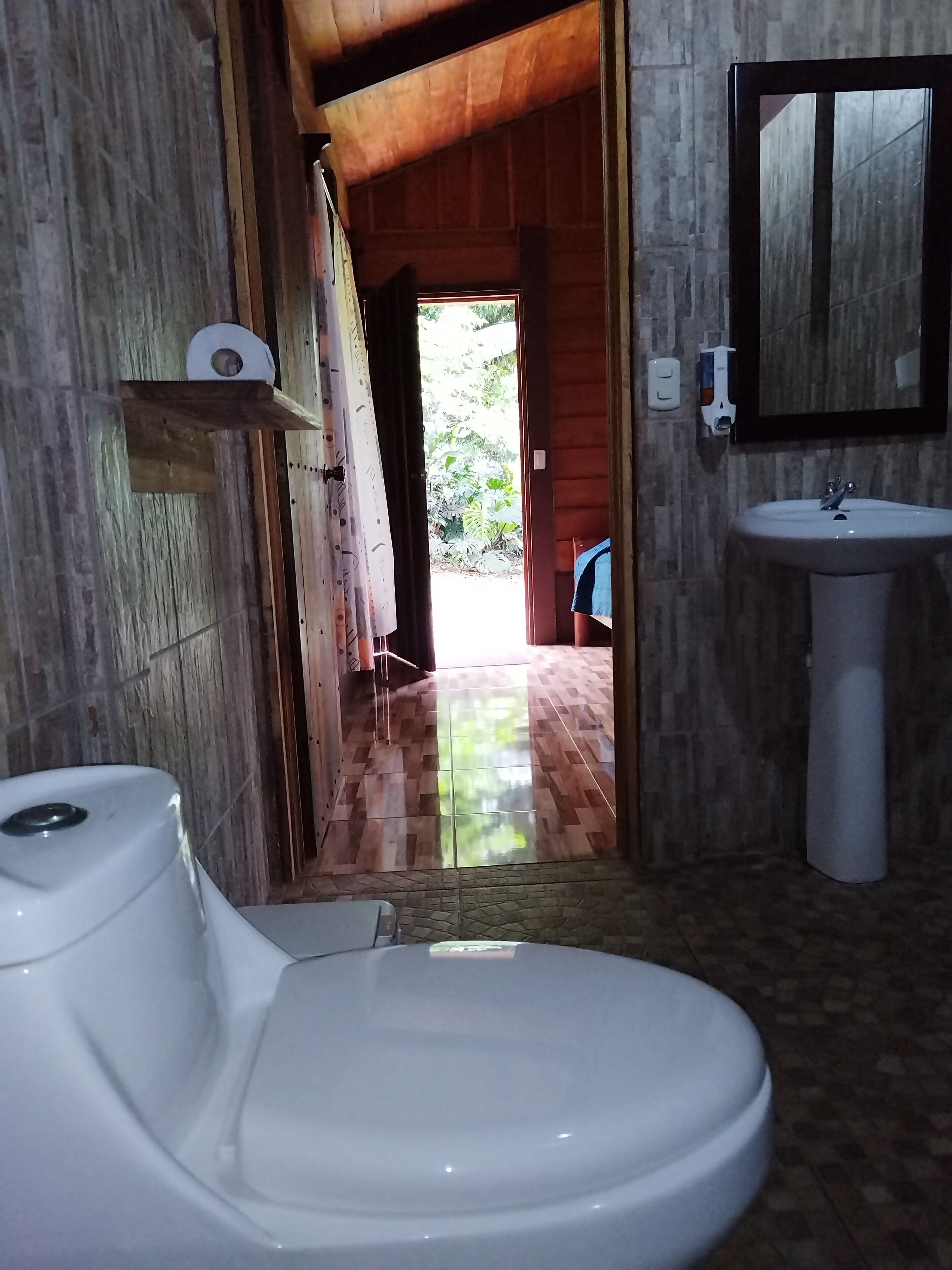 A bathroom with a toilet, sink, and shower area. There is a wooden door leading to an outdoor area with a tiled floor and greenery.