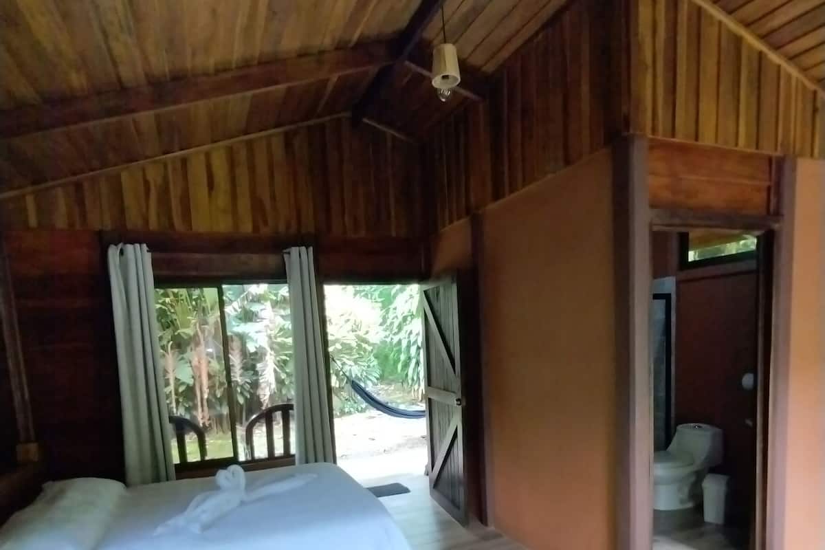 A wooden room with a bed, a door leading to a bathroom, and a view of greenery outside.