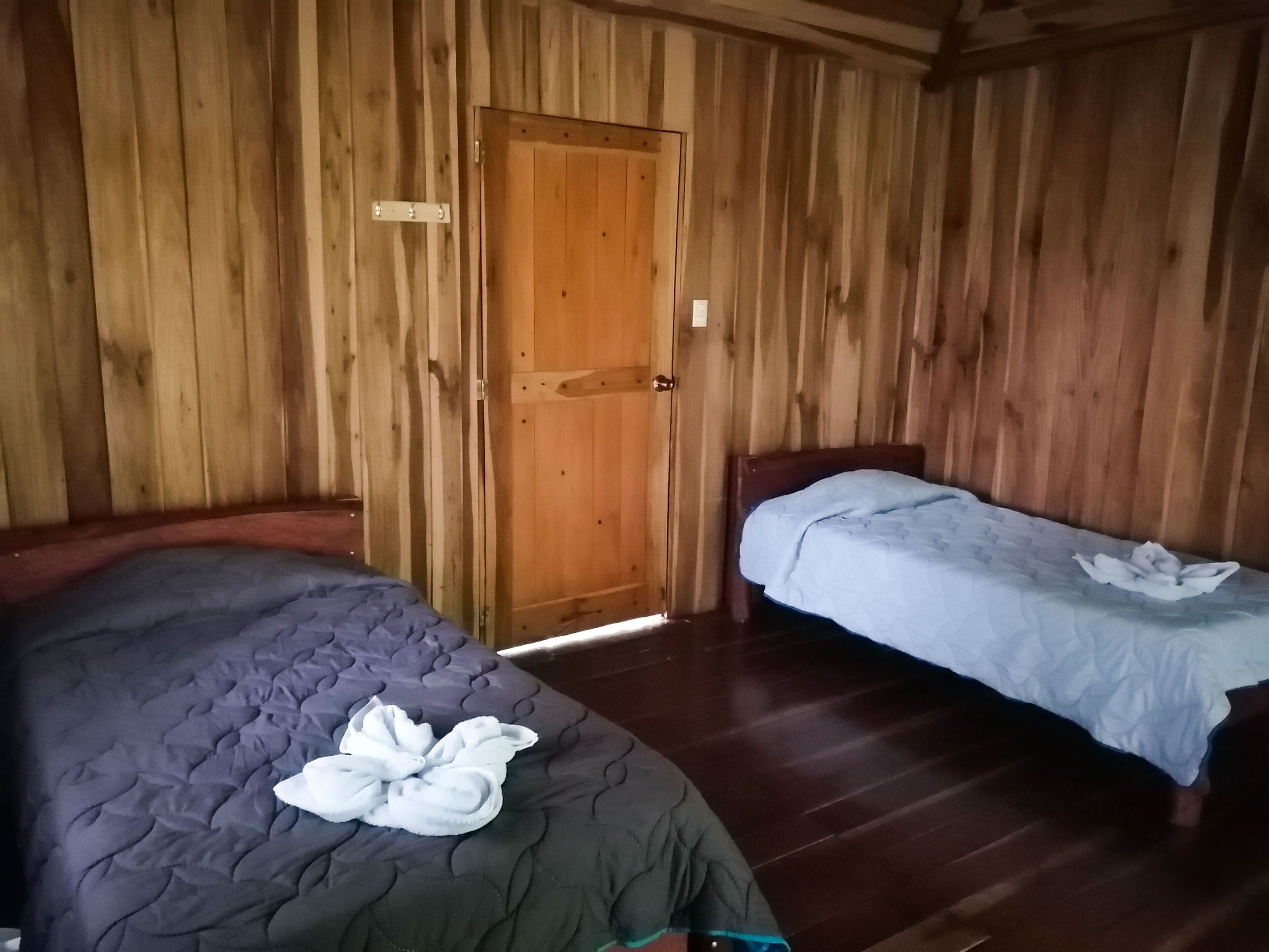 Une pièce aux murs en bois, avec une porte, un lit recouvert d’une couverture sombre et un plus petit lit avec une couverture de couleur claire.