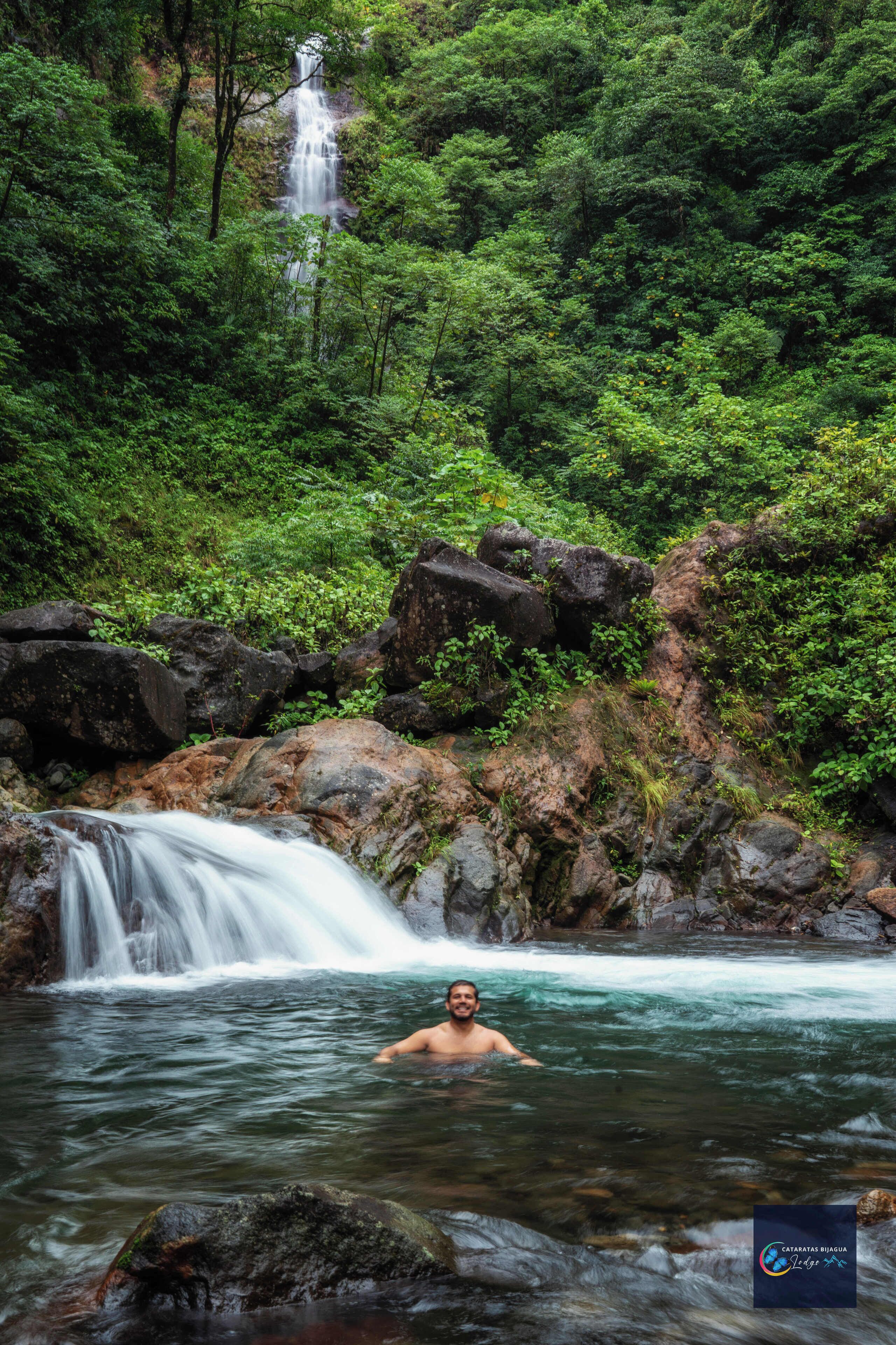 A man in a natural setting with a waterfall and lush greenery.