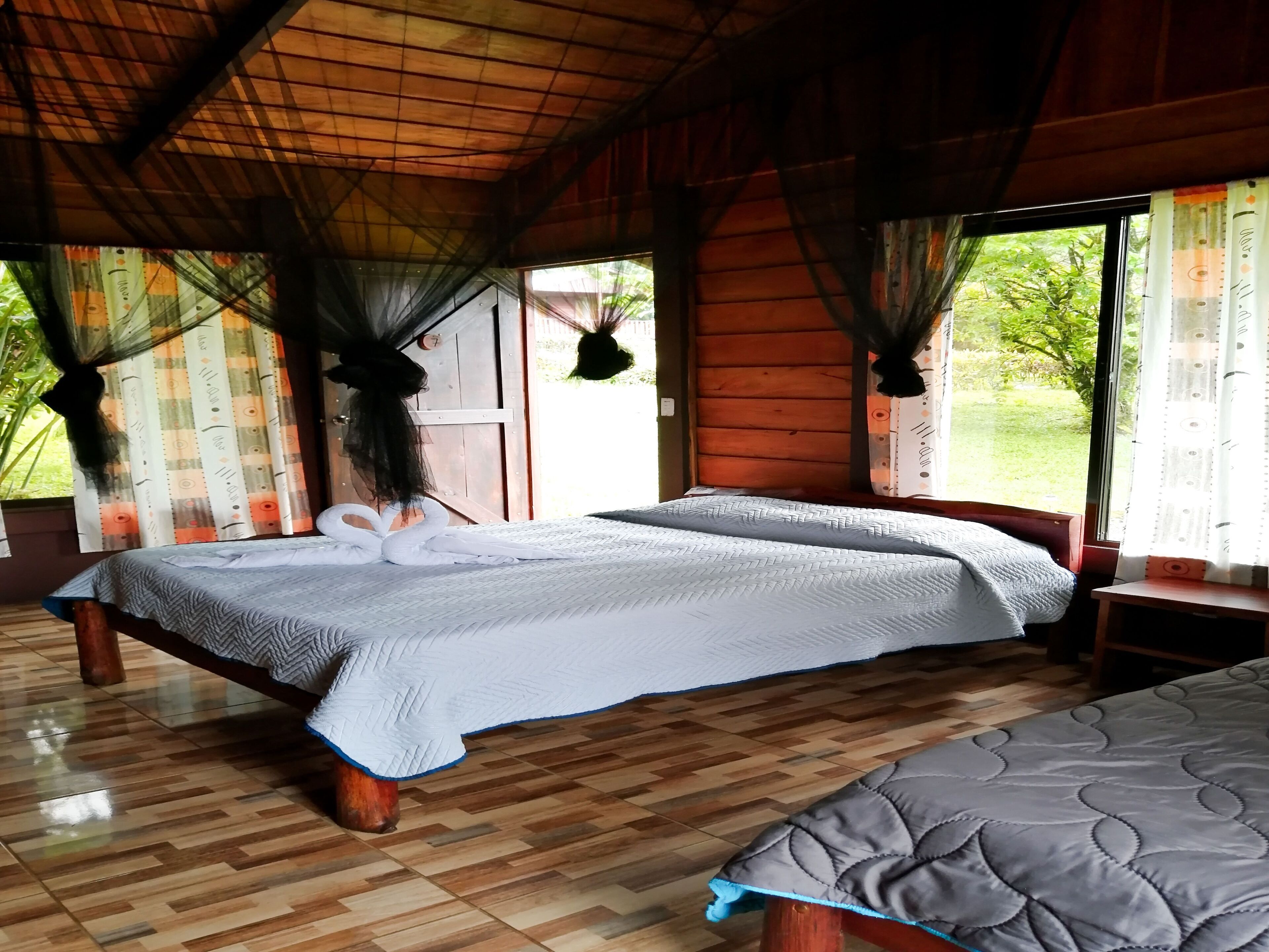 A room with two beds, wooden flooring, and large windows with curtains.