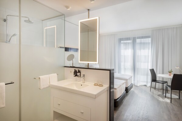 Quarto de hotel moderno com um espelho grande, toucador branco com lavatório, cama, mesa pequena com cadeira e planta em vaso.