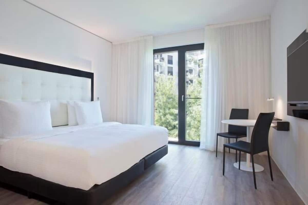 Une chambre d’hôtel avec un grand lit, une petite table avec deux chaises et une vue sur la verdure par la fenêtre.
