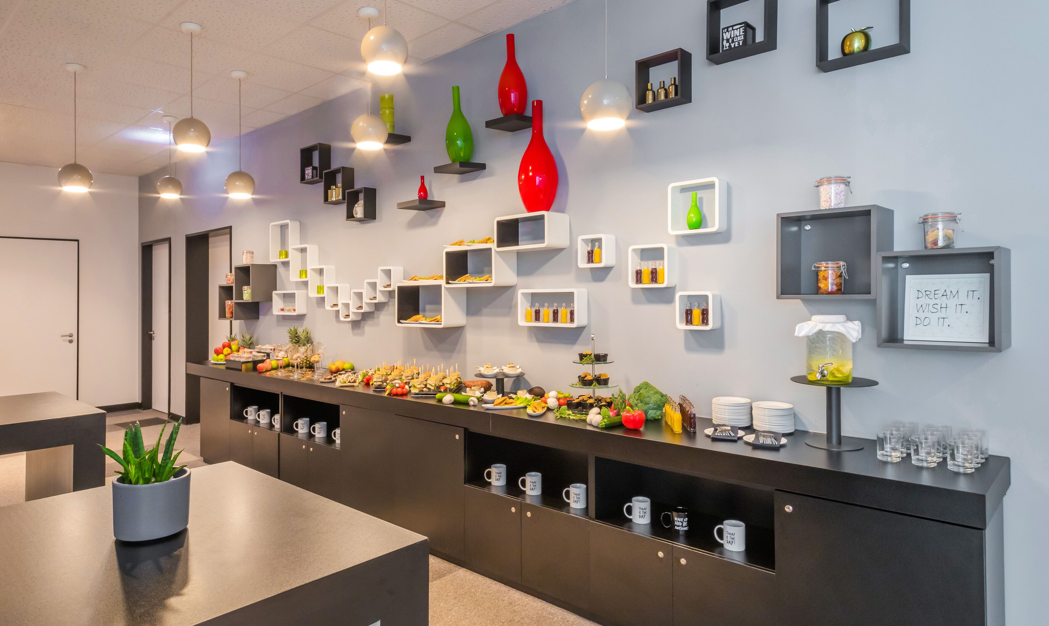 A modern kitchen with a long counter, various food items, and a wall with shelves displaying bottles and a motivational quote.