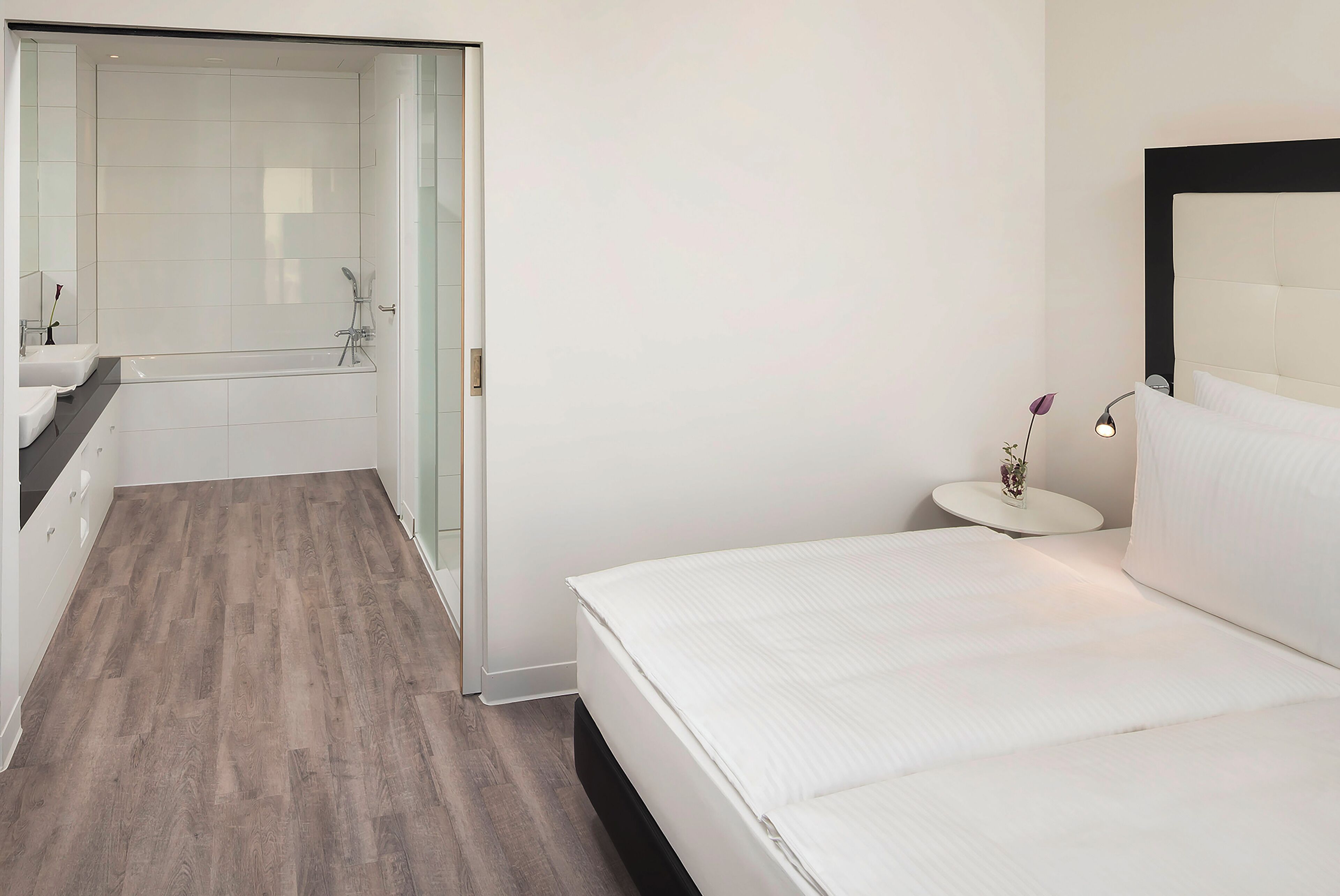 Una habitación de hotel moderna con una cama grande, un baño independiente con bañera y una mesita de noche pequeña con lámpara.