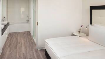 Una habitación de hotel moderna con una cama grande, un baño independiente con bañera y una mesita de noche pequeña con lámpara.
