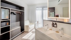 A modern hotel room with a walk-in closet, a large mirror, a white sofa, a small round table with a plant, and a bathroom with a double sink and vanity. - INNSiDE by Meliá Frankfurt Ostend (Frankfurt)