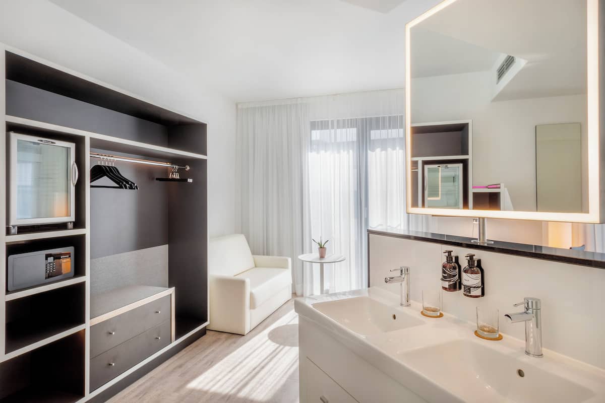 Une chambre d’hôtel moderne dotée d’un dressing, d’un grand miroir, d’un canapé blanc, d’une petite table ronde agrémentée d’une plante, et d’une salle de bain avec un double lavabo et un meuble-vasque.