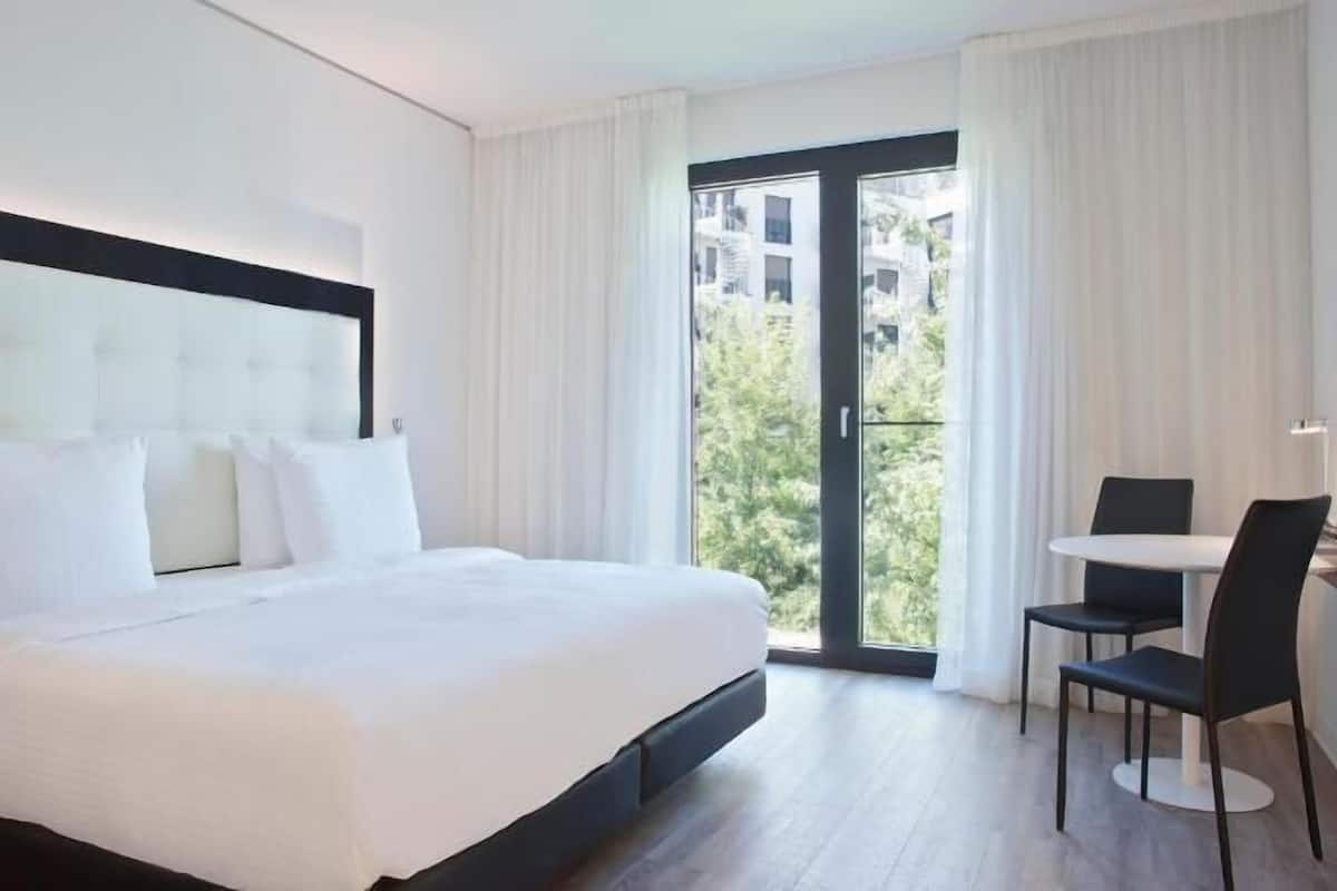 Une chambre d’hôtel avec un lit, une petite table et des chaises, une télévision et un balcon avec des rideaux.