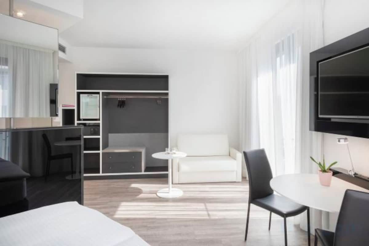 Una habitación de hotel moderna con una cama, un escritorio, una silla, una mesita, un sofá, un televisor y un armario.