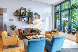 Una sala de reuniones moderna con un sofá de cuero marrón, sillones azules y una mesa de centro. Hay plantas en macetas sobre estantes y un ventanal con vistas a árboles.