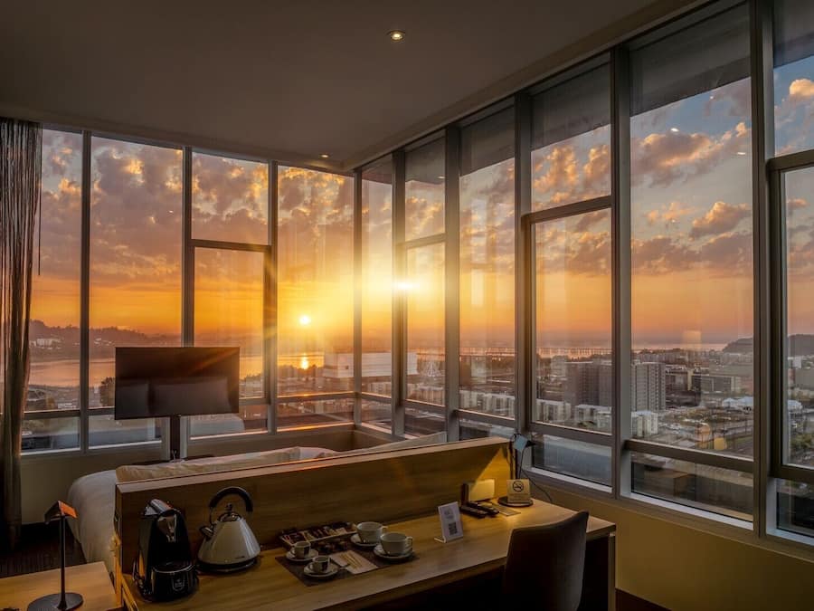 Ein Hotelzimmer mit großem Fenster und Blick auf die Stadt, einem Schreibtisch mit Wasserkocher und einem Bett.
