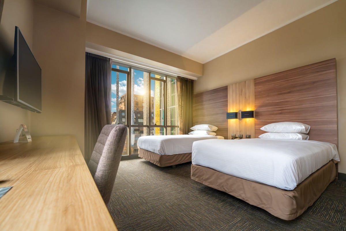A hotel room with a desk, two beds, a TV, and a large window with curtains.