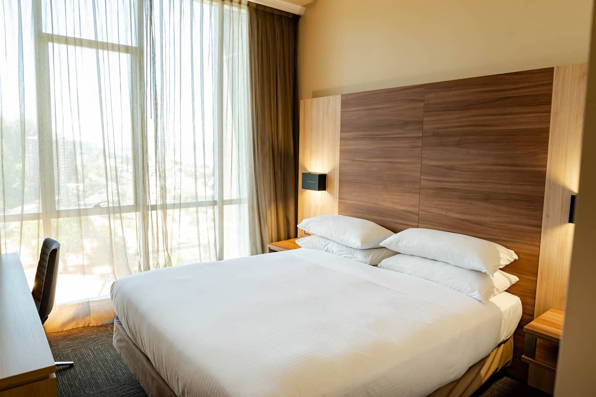 A hotel room with a large bed, a desk, and a chair. There is a window with curtains and a view of the outside.