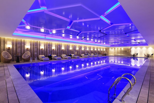 Piscina interior moderna com iluminação azul, cadeiras de descanso e design de teto geométrico.